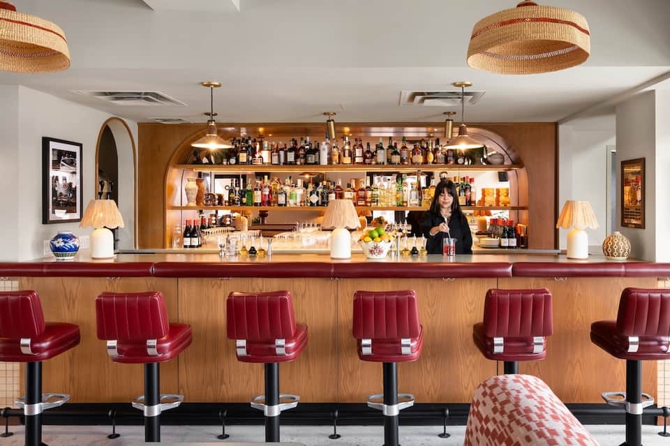 Hotel bar with wood-paneled back wall, burgundy bar stools, shelves of spirits, and warm pendant lighting
