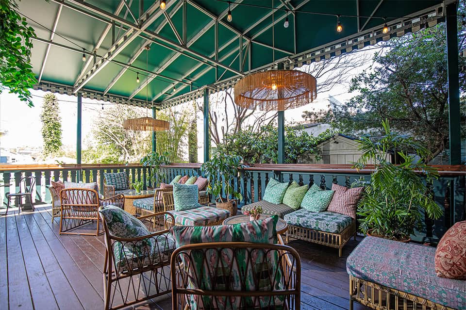 Covered outdoor patio with rattan furniture, colorful cushions, and lush green plants