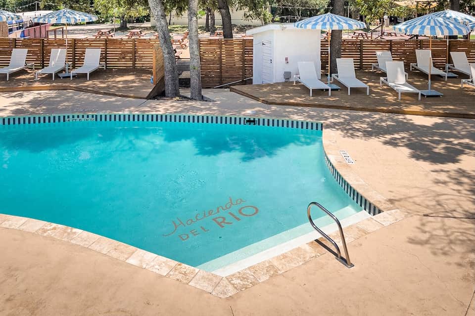 Swimming pool with 'Hacienda Del Rio' text, white lounge chairs, blue umbrellas, and wooden fence