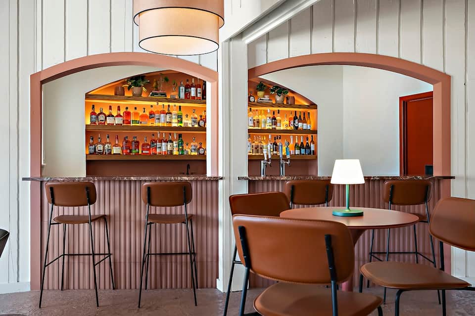 Modern bar lounge with two bar stations, extensive liquor shelves, brown leather stools, and contemporary lighting
