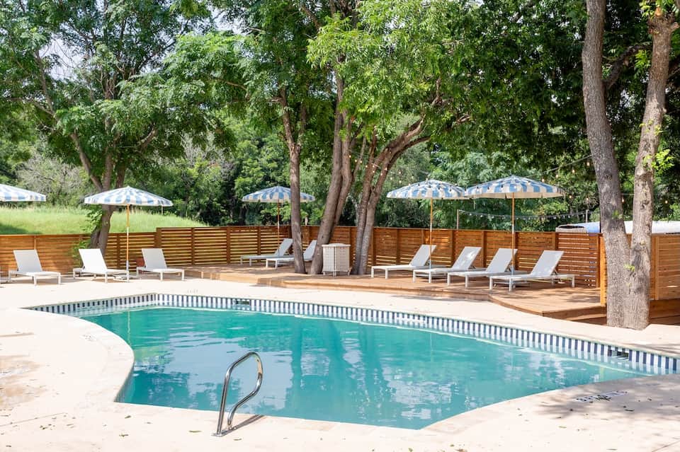 Resort pool with striped umbrellas, lounge chairs, wooden deck, and tall shade trees