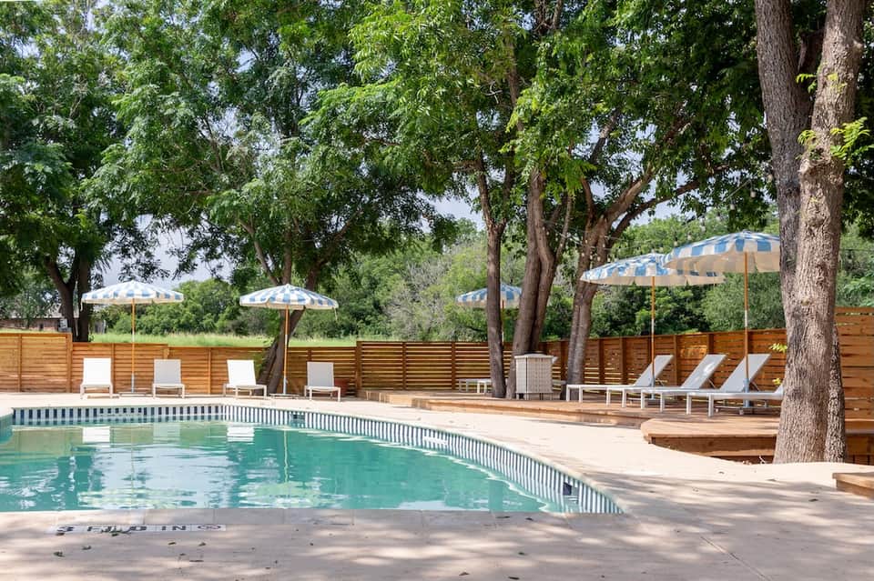 Poolside lounge under mature trees with striped umbrellas, white loungers, and wooden privacy fence
