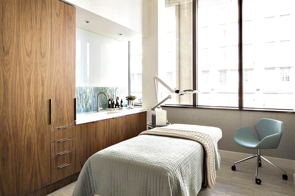 Modern spa treatment room with wooden cabinetry, white marble countertop, and floor-to-ceiling windows overlooking city