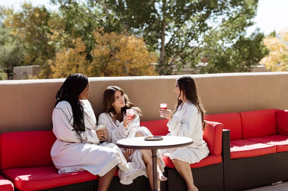 Three women in spa robes enjoying beverages on orange patio seating with ocean view