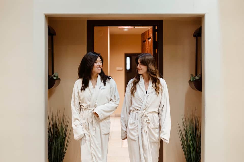 Two women in white robes walking through a spa hallway corridor