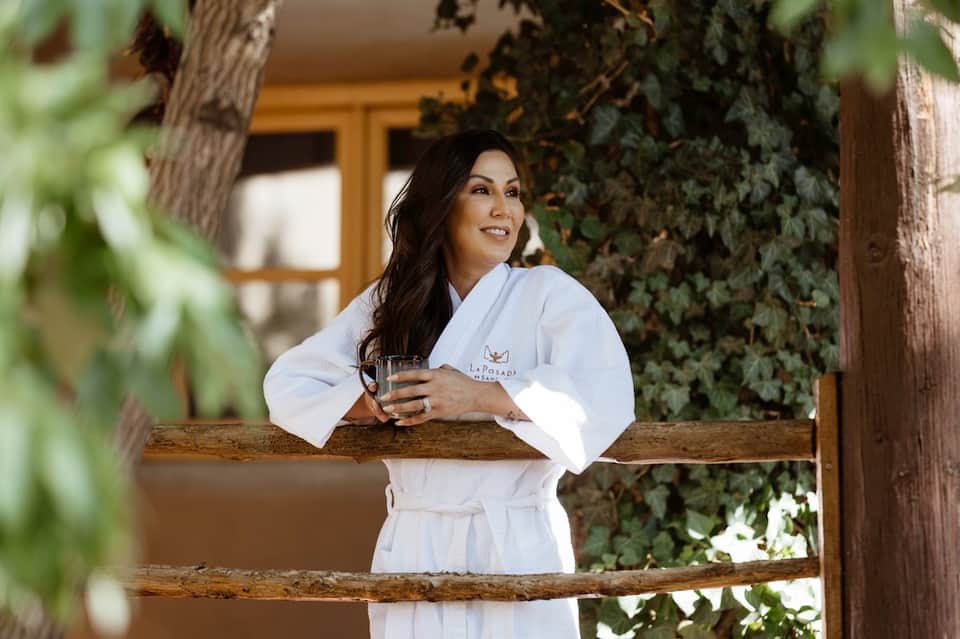 Woman in white spa robe holding a beverage on a wooden deck surrounded by ivy and greenery