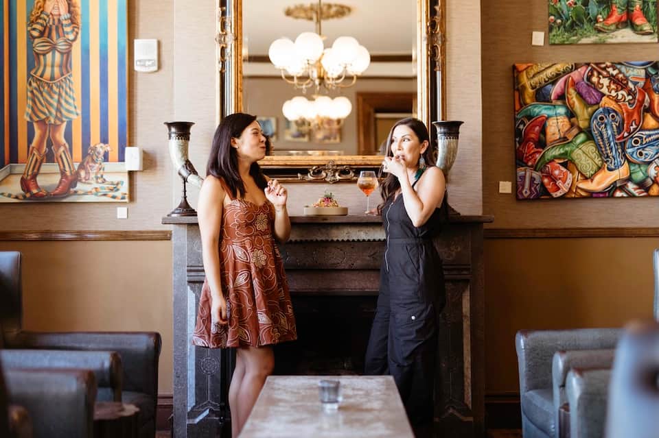 Two women at elegant bar interior with gold-framed mirror, artwork, and brass candlesticks