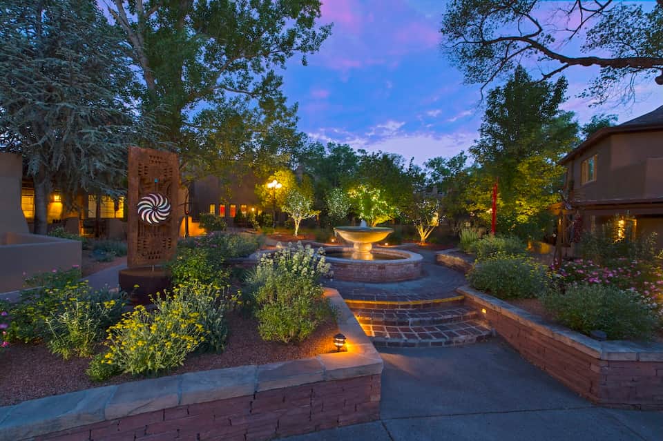 Illuminated courtyard at dusk with fountain, decorative wall art, landscaping, and building lights