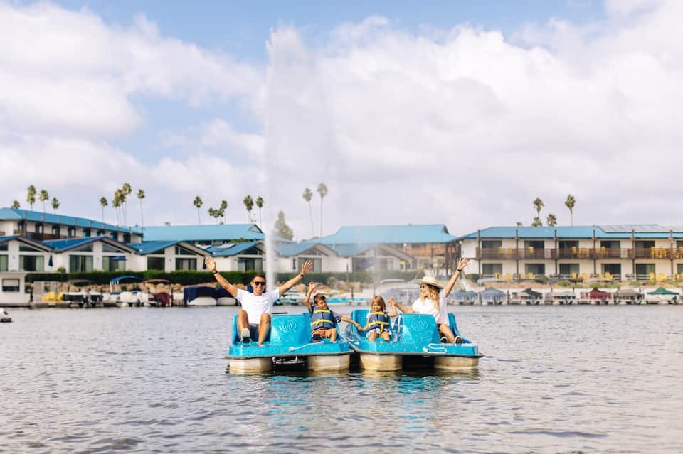Family enjoying bright blue pedal boat on water with coastal buildings and palm trees in background