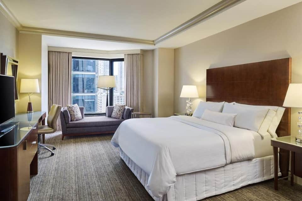 Luxury hotel room with king bed, sofa, city skyline views, and warm lighting