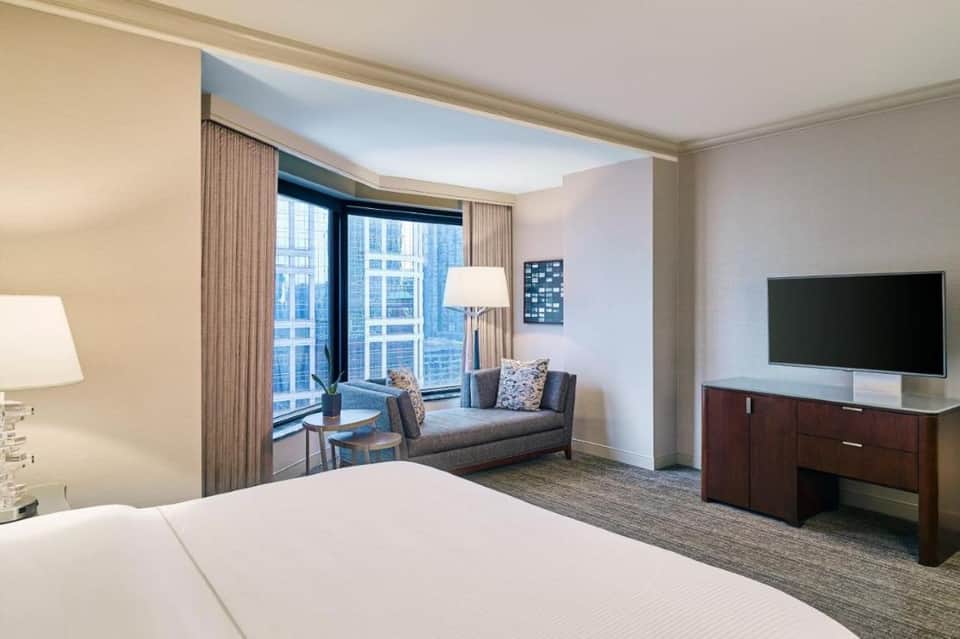 Modern hotel bedroom with king bed, sofa seating area, and floor-to-ceiling windows
