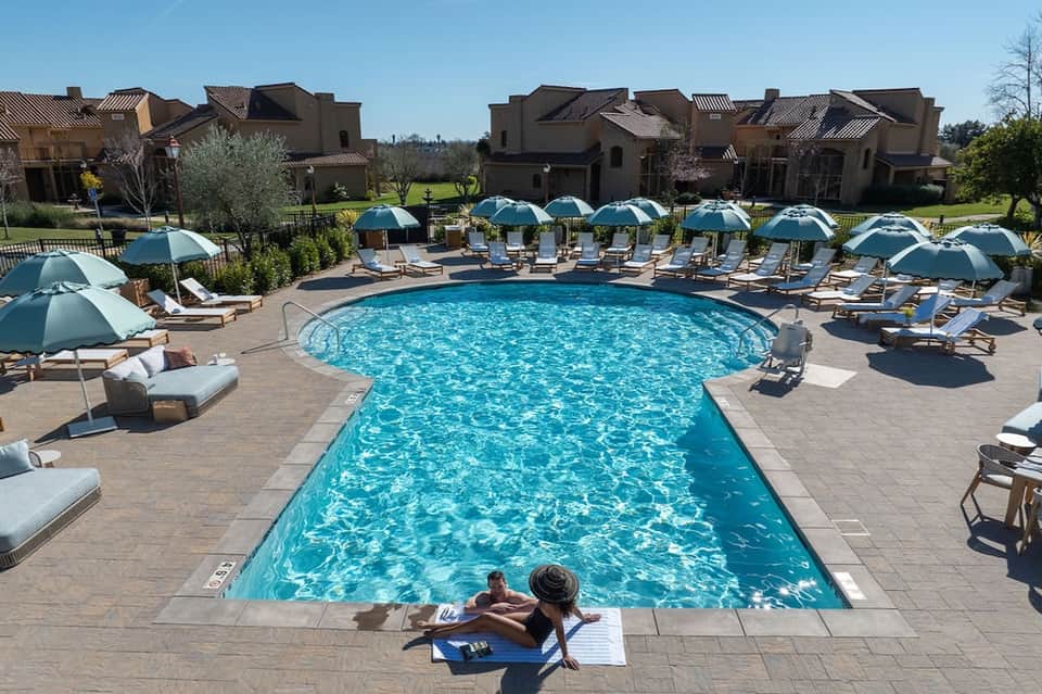 Curved resort pool surrounded by blue umbrellas, lounge chairs, and Mediterranean-style villa buildings