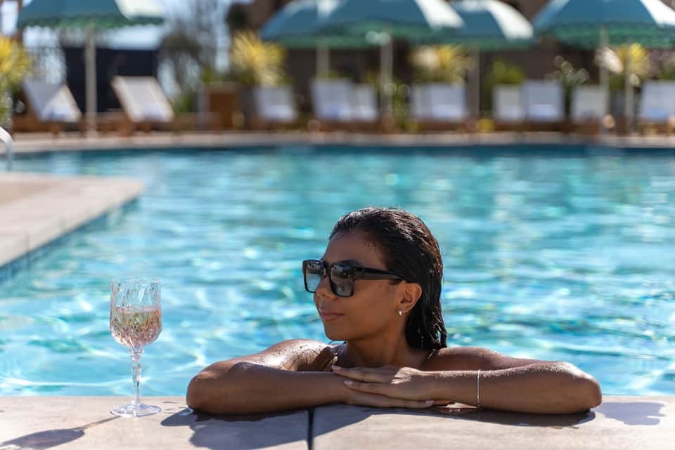 Woman in sunglasses relaxing in resort pool with wine glass, beach umbrellas visible in background