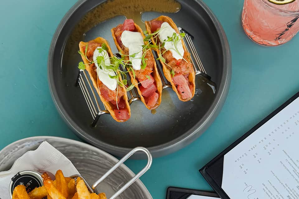 Gourmet meal of three colorful tacos on a black plate with a pink cocktail on turquoise table