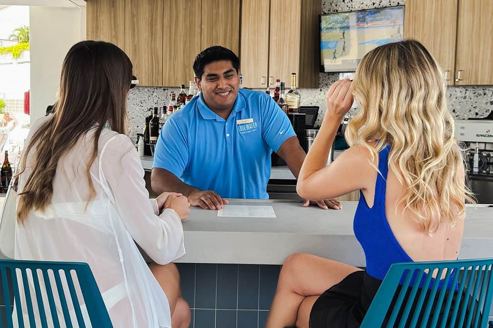 Bartender in blue polo greeting two female guests at resort bar counter