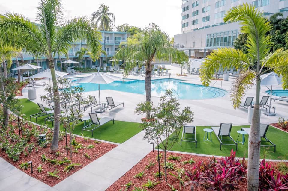 Resort pool area with palm trees, lounge chairs, umbrellas, and tropical landscaping