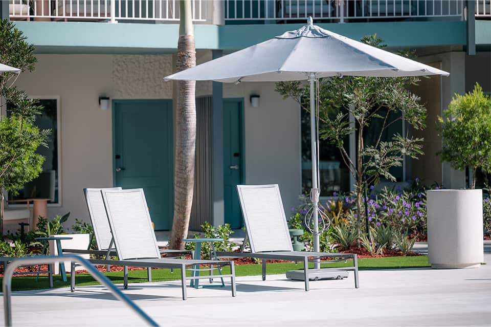 Resort patio with white lounge chairs, gray umbrella, palm tree, and teal building entrance