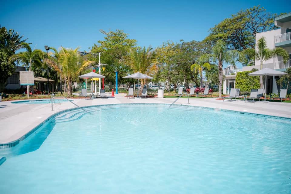 Family-friendly resort pool with palm trees, white umbrellas, and colorful children's play area