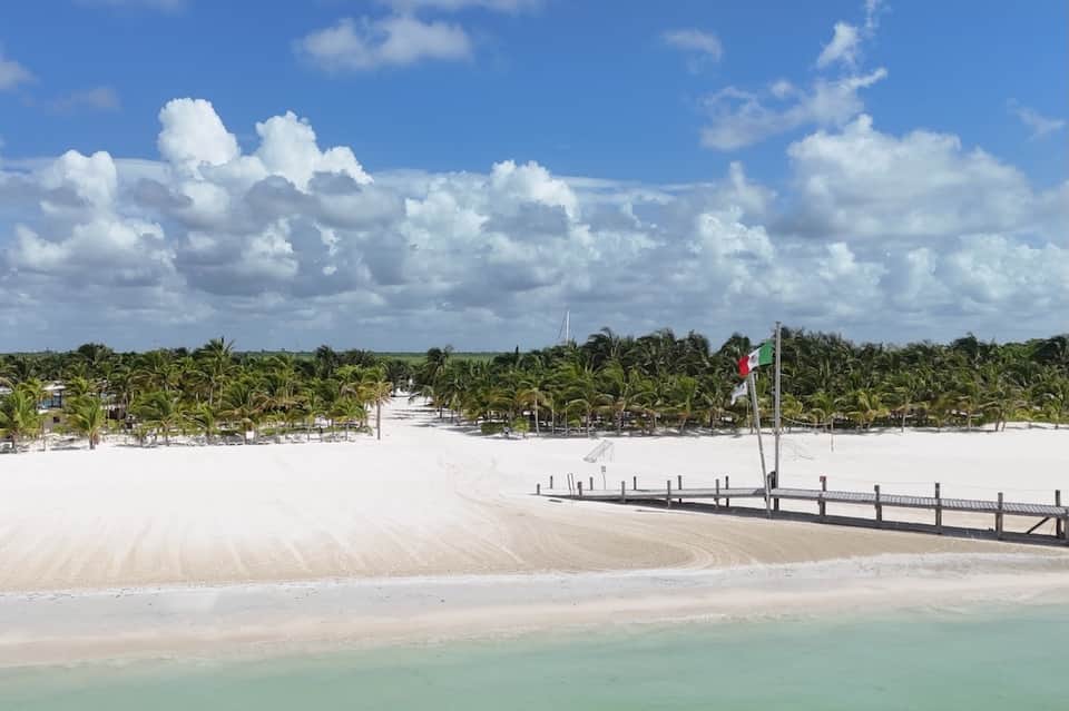 Pristine beach with white sand, turquoise water, palm trees, and wooden dock with Mexican flag