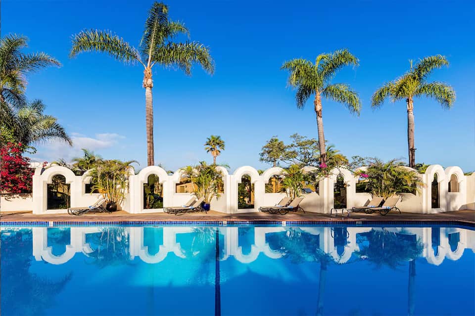 Resort pool with white arched cabanas, palm trees, red flowers, and clear blue water under sunny sky