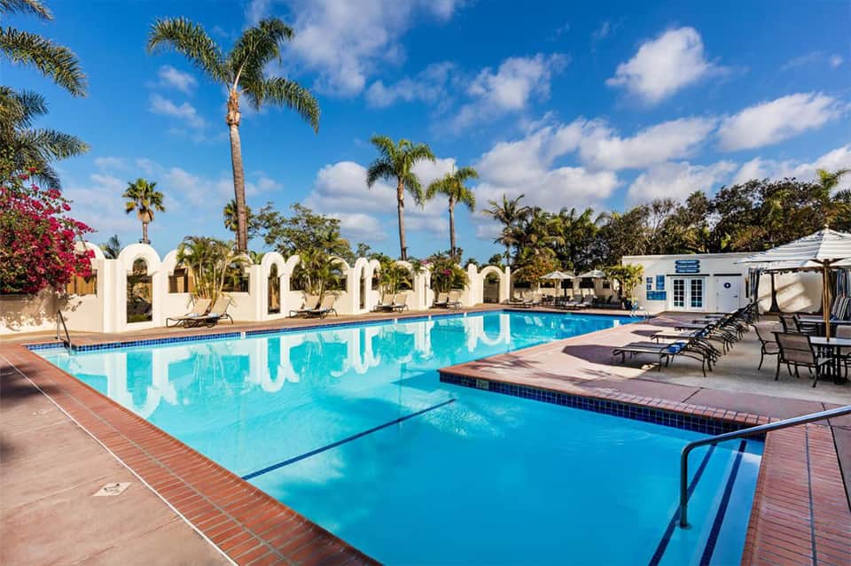 Large turquoise swimming pool with white arched structures, palm trees, and lounging areas under clear sky