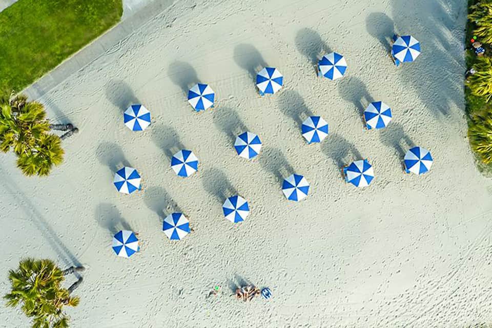 Aerial view of sandy beach with blue and white striped umbrellas arranged in rows and palm trees