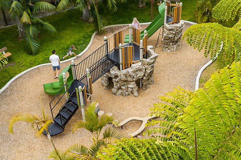 Aerial view of playground with green slides, wooden play equipment, safety fencing, grass, and palm trees