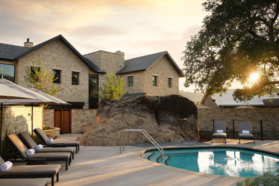 Luxury resort pool at sunset with loungers, stone house, large rock feature, and mature trees