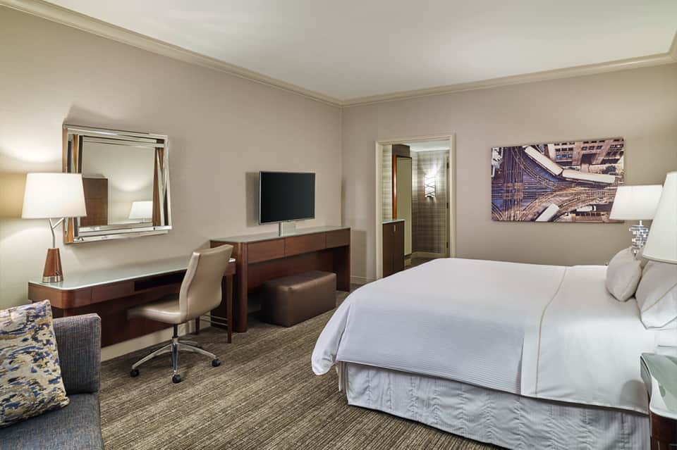 Hotel bedroom with king bed, work desk, flat-screen TV, and city artwork on beige walls