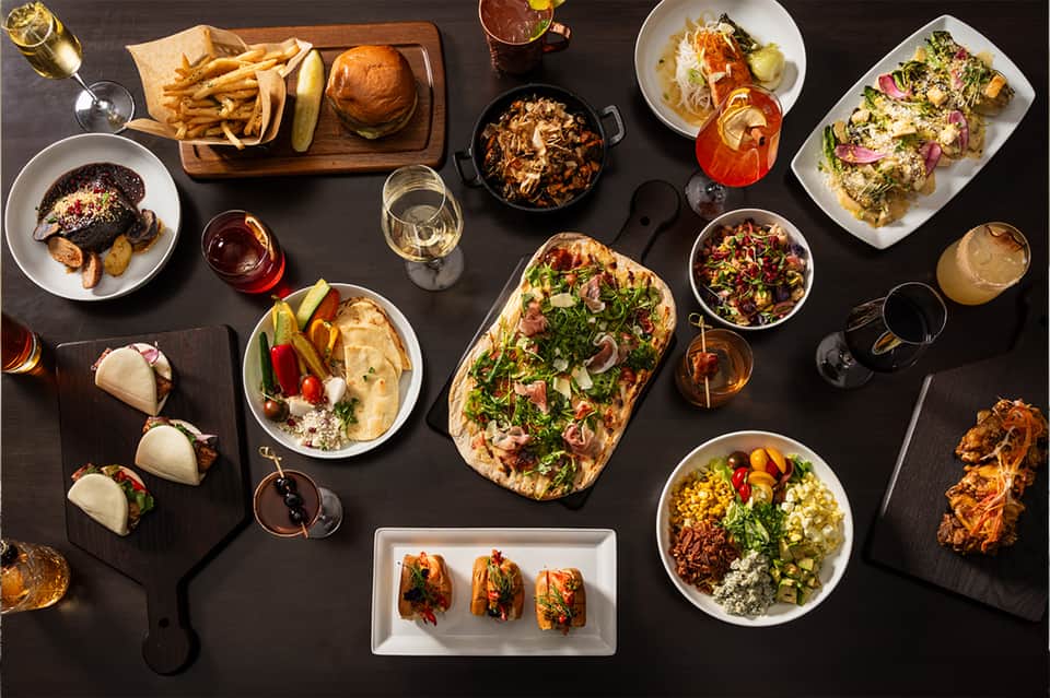 Resort dining spread with tacos, burgers, seafood dishes, cocktails, and appetizers on black table