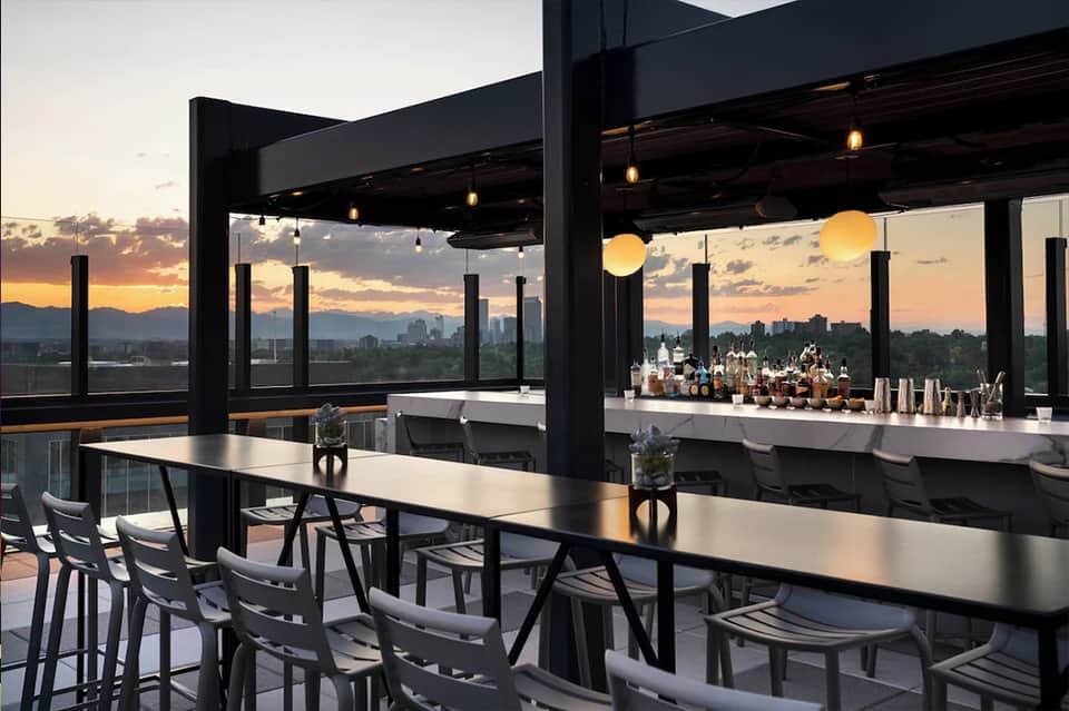 Rooftop bar at sunset with modern black structure, illuminated bar, city skyline, and mountain views