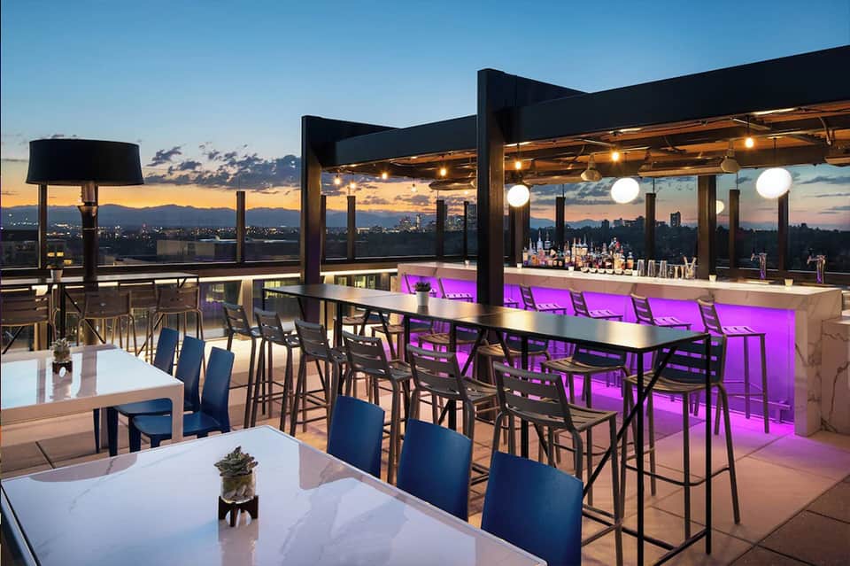 Rooftop bar at dusk with purple lighting, modern seating, and panoramic city skyline views