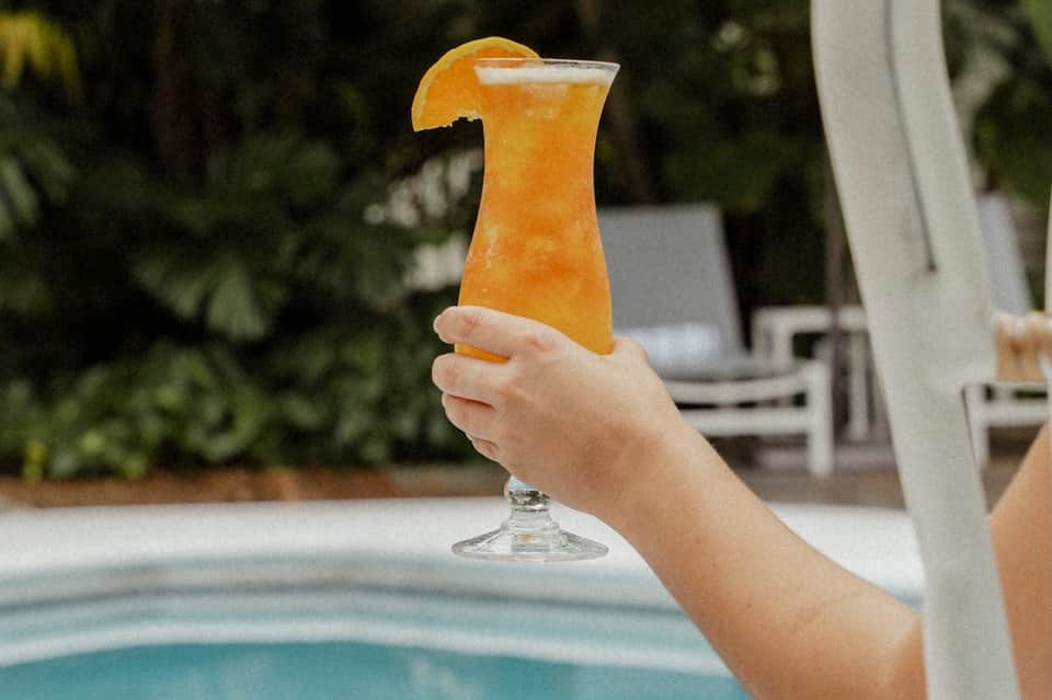Hand holding tropical orange cocktail with orange slice garnish by resort pool