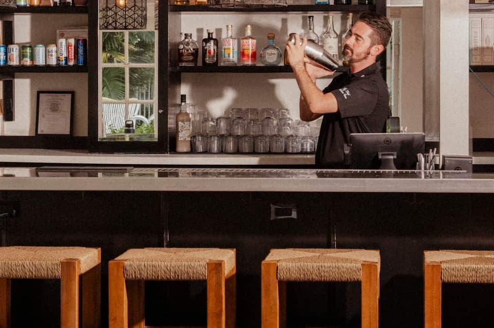 Bartender in black shirt mixing cocktail behind modern bar with shelves of spirits and glassware