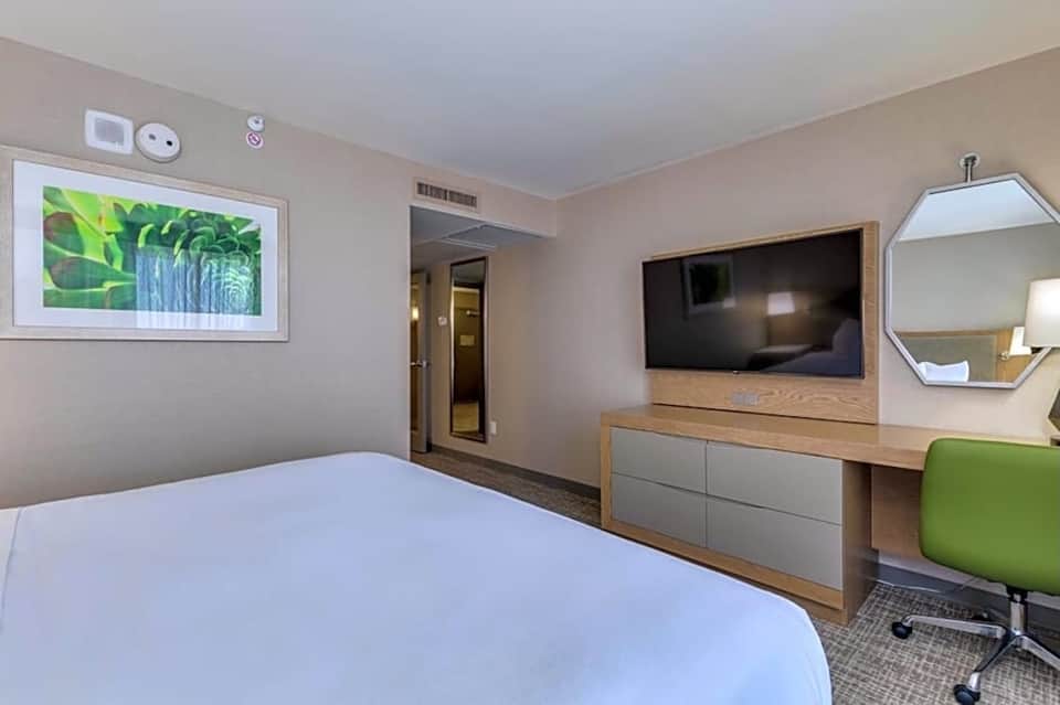 Hotel room with queen bed, TV mounted above wooden dresser, green chair, and digital art display