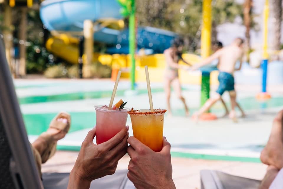 Two colorful cocktails with straws held up by poolside with water park features and guests in background