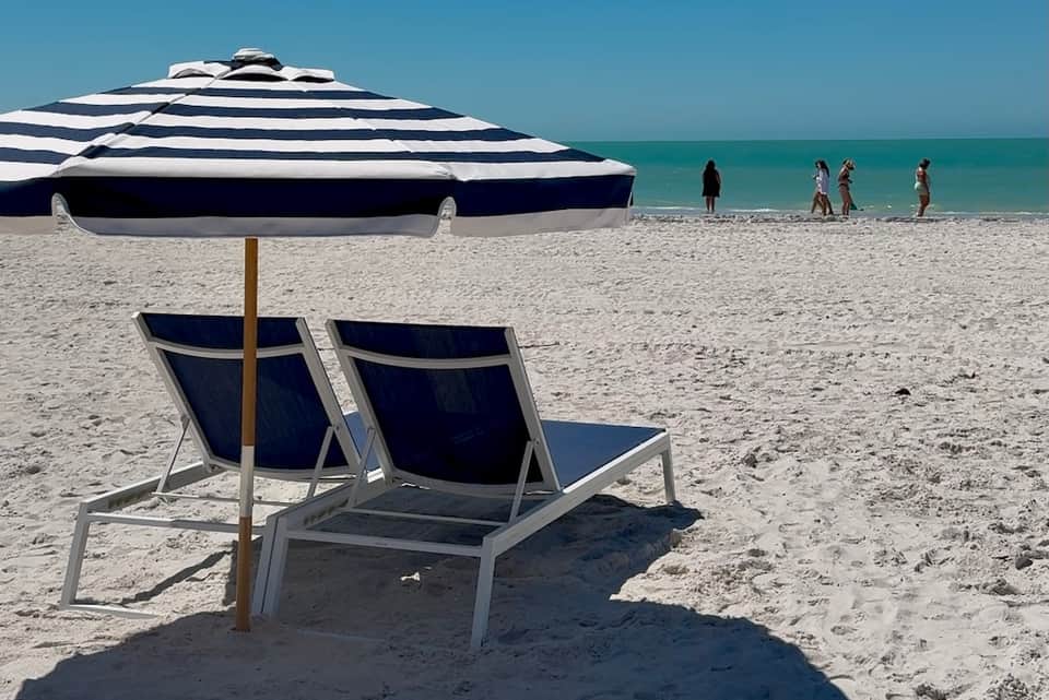Beach scene with navy and white striped umbrella, lounge chairs on sand, ocean, and beachgoers in distance