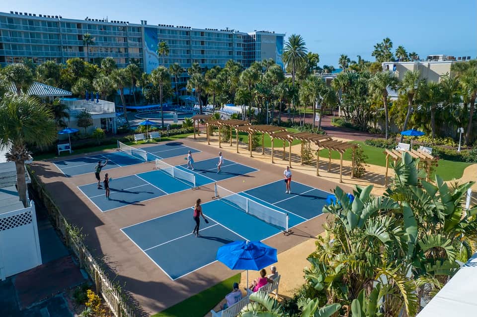 Aerial view of multiple pickleball courts with players, palm trees, resort building, and manicured grass areas