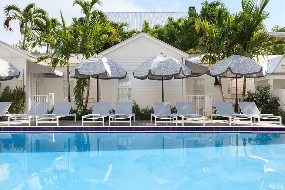 Resort pool with blue water, lounge chairs, patterned umbrellas, white cottages, and palm trees