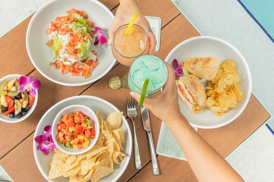 Poolside dining with fresh salads, sandwiches, fruit bowls, and colorful tropical drinks