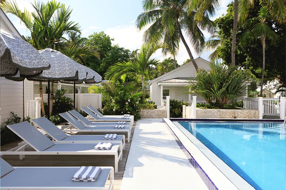 Resort pool deck with white lounge chairs, thatched umbrella, palm trees, and cottage-style buildings