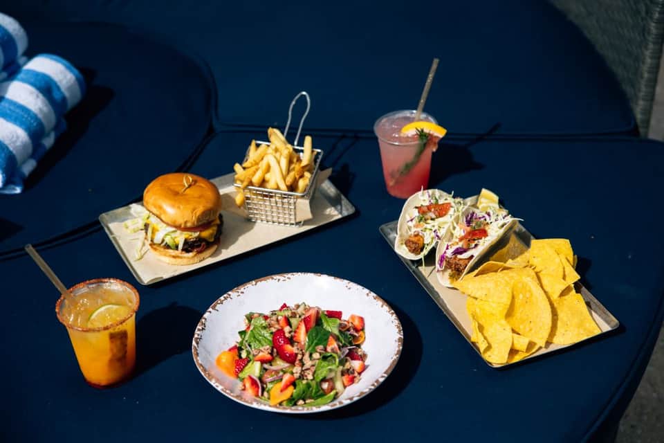 Poolside food and drinks including burger, fries, tacos, salad, and colorful cocktails on blue table