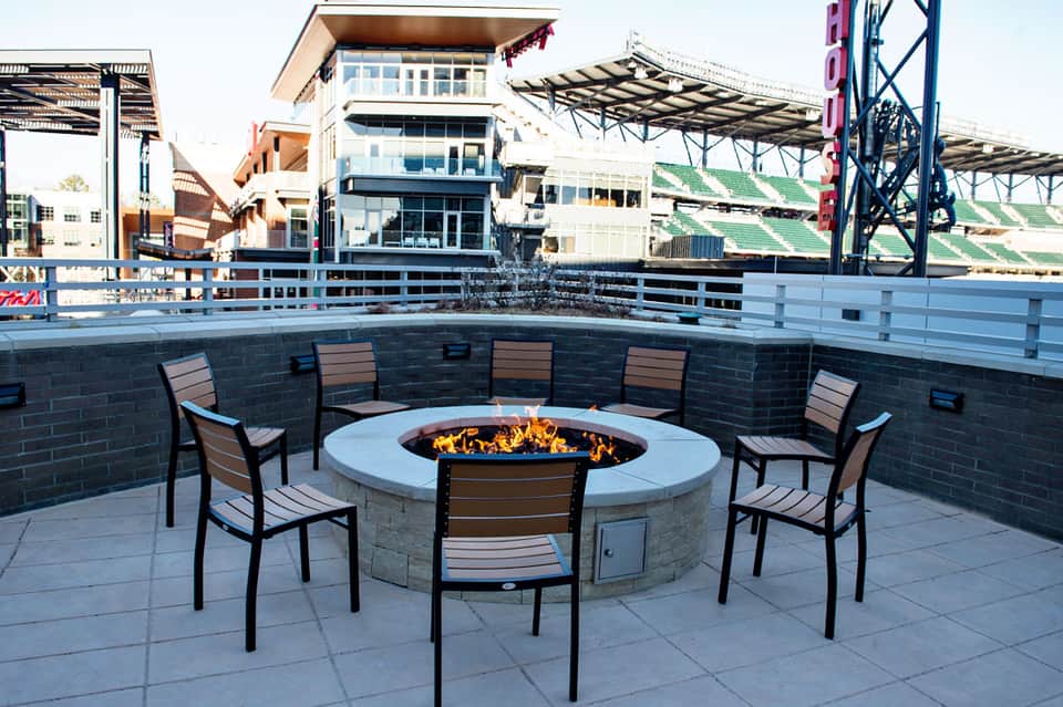 Modern outdoor fire pit lounge area with seating surrounded by sports stadium and buildings