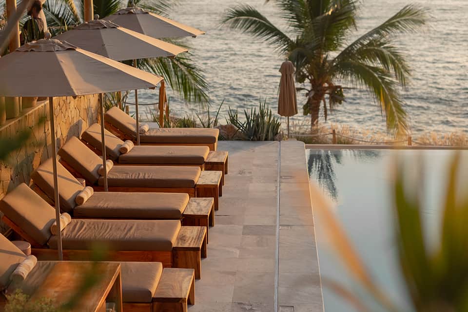 Beachfront lounge area with cushioned sunbeds under umbrellas, palm trees, and ocean view at golden hour