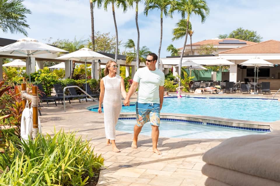 Couple walking along resort pool deck with palm trees, umbrellas, loungers, and tropical landscaping