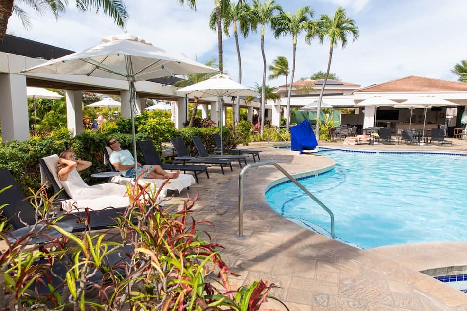Resort poolside with lounge chairs under umbrellas, palm trees, and tropical landscaping on sunny day