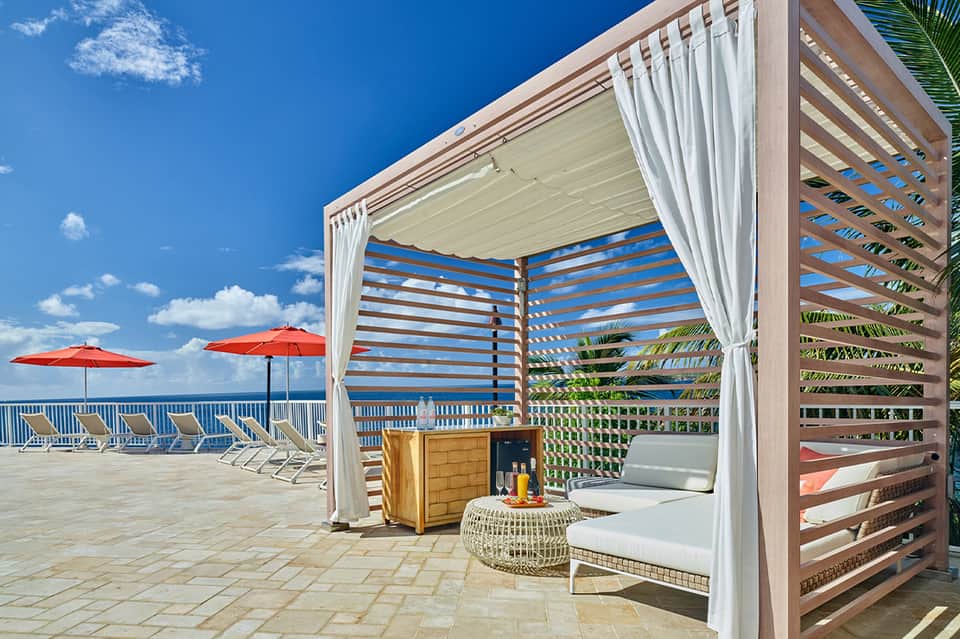 Oceanfront cabana with retractable roof, white curtains, red umbrellas, and lounge seating