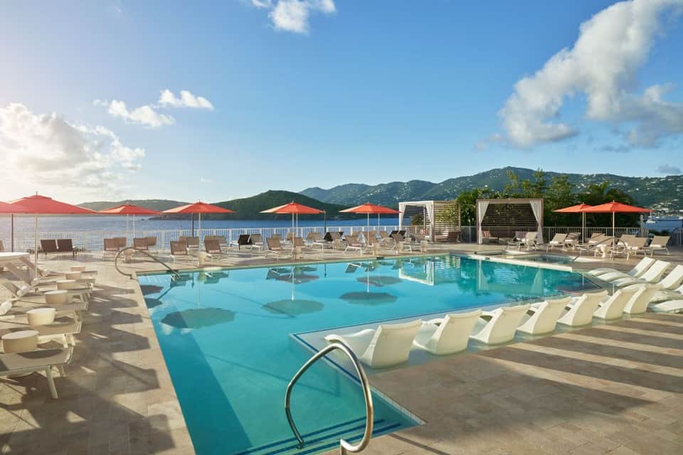 Resort pool with turquoise water, lounge chairs, red umbrellas, and mountainous waterfront views