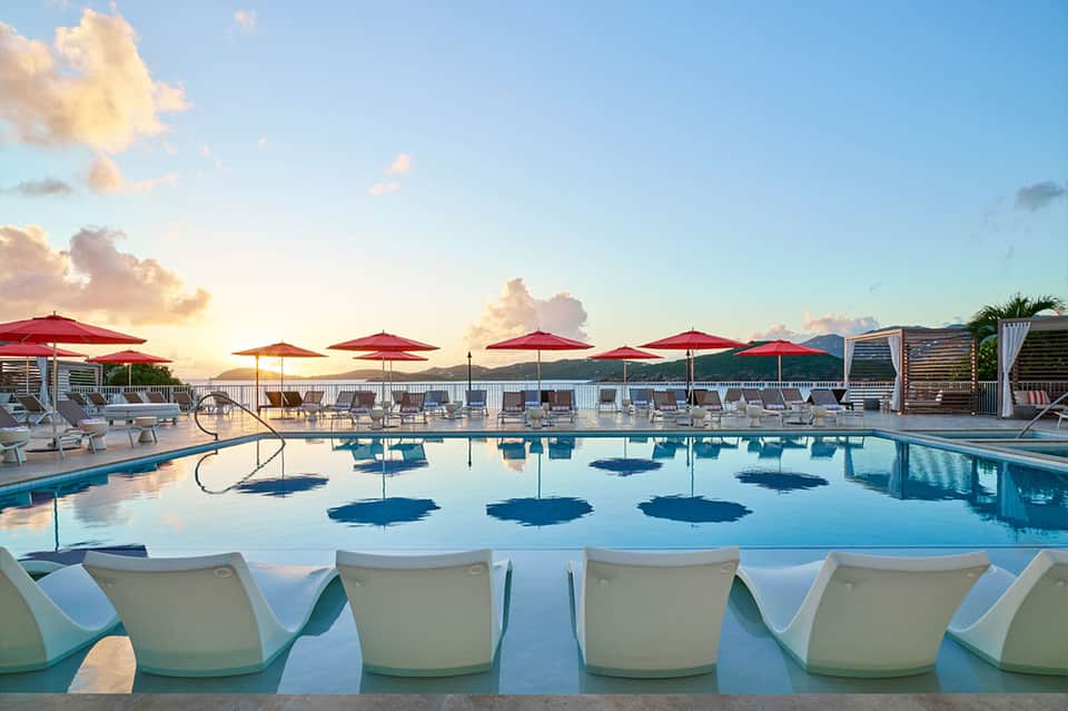 Sunset pool scene with red umbrellas, white lounge chairs, and mountains in background