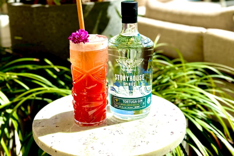 Colorful cocktail with pink flower garnish next to a bottle of Storyhouse gin on outdoor table